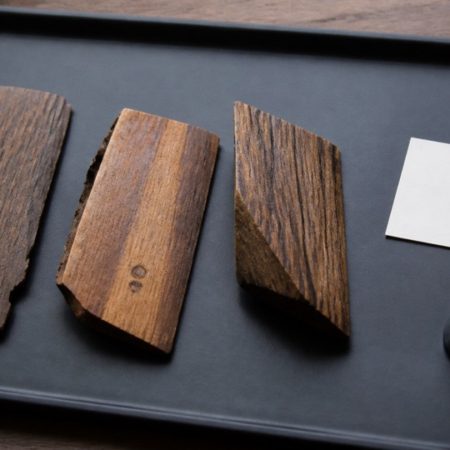 Unlabelled natural wood pieces on a dark tray with small circular surface marks, a blank card and a magnifying loupe
